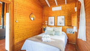 A wooden cabin bedroom with a bed, a round mirror, framed artwork, and a small shelf with decorative items. - Hornillos Alozaina by Ruralidays (Málaga)