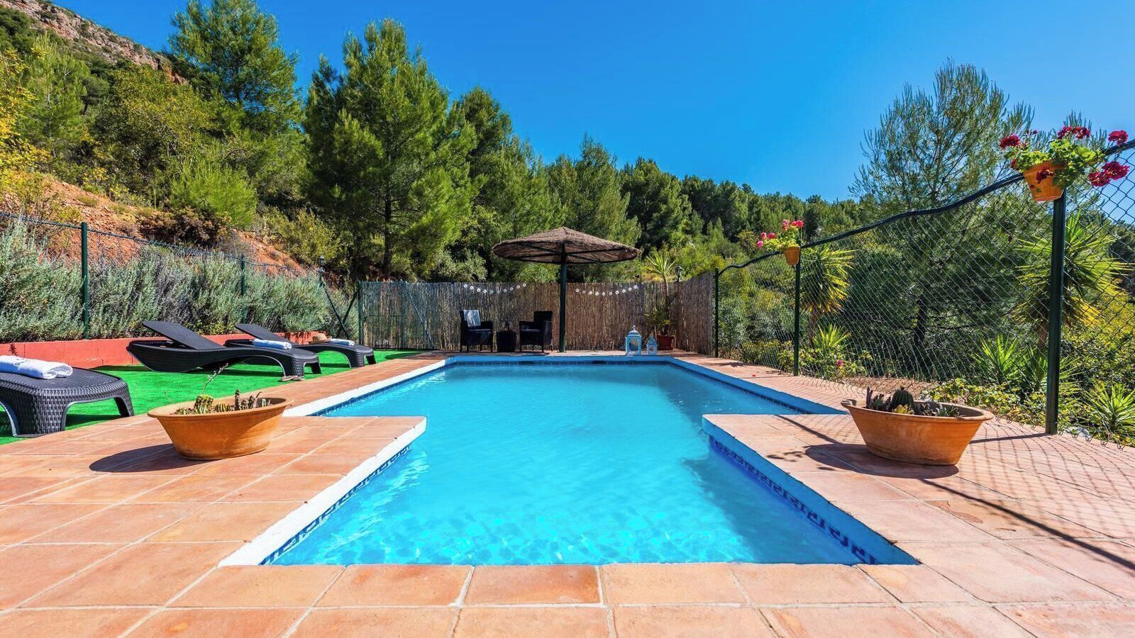 Ein rechteckiger Pool mit einer gefliesten Kante, umgeben von einer Pflasterterrasse. Im Hintergrund sind Liegestühle und eine kleine Sonnenschirmkonstruktion zu sehen.