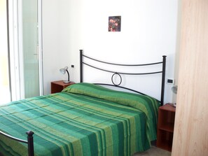 A single bed with a green striped bedspread, a bedside table with a lamp, and a small picture on the wall. - Appartamenti Pigafetta (Valledoria)