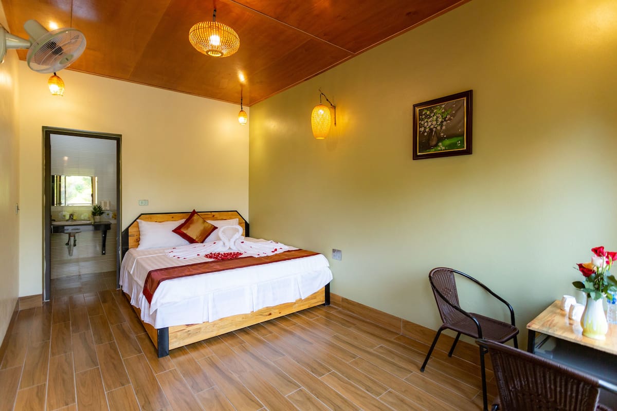 A hotel room with a wooden bed, a dining table with chairs, a wall-mounted fan, and a painting.