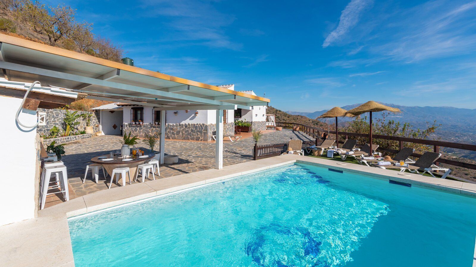 A pool area with a covered seating area, lounge chairs, and a view of the mountains.