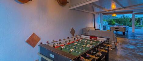 A foosball table in a room with a pool table and a covered outdoor area.