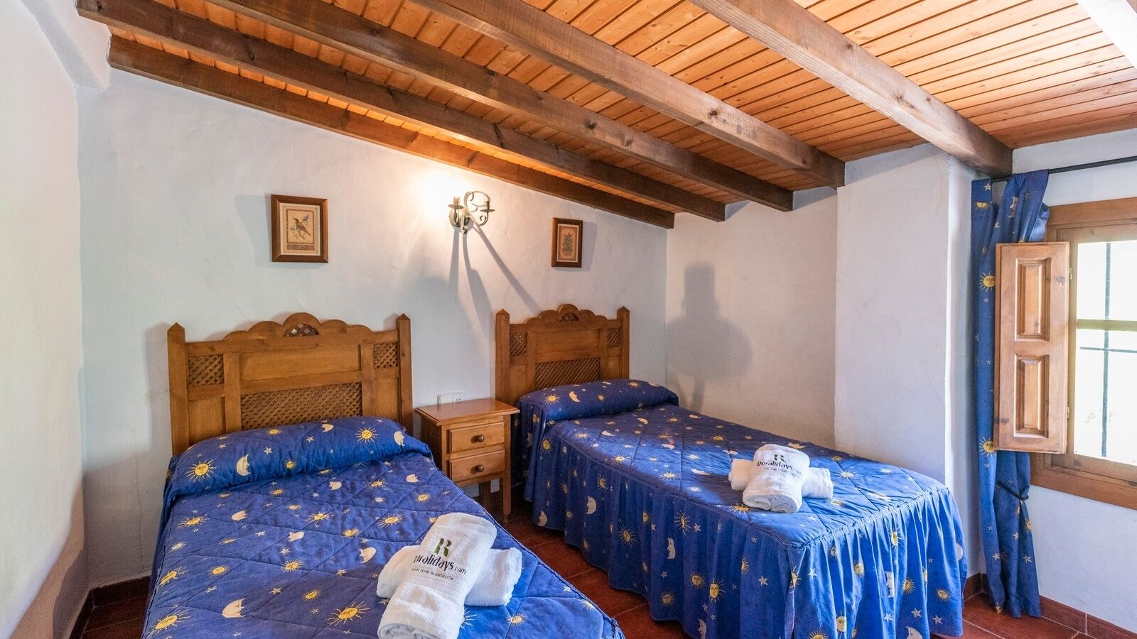 A room with two wooden beds, each with a blue bedspread featuring a sun and moon pattern. There are two wall-mounted lamps and a wooden cabinet with a window.