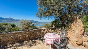 Une terrasse en pierre avec une table et des chaises, offrant une vue panoramique sur un paysage magnifique, avec des montagnes et une étendue d’eau.