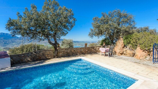 Une piscine bordée d’un mur en pierre et une table avec des chaises, le tout entouré d’arbres et offrant une vue sur les montagnes.