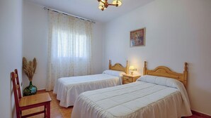 Ein Zimmer mit zwei Einzelbetten, einem Nachttisch aus Holz mit einer Lampe, einem Sessel und einem Fenster mit Vorhängen.
