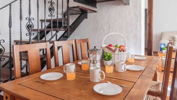 A wooden dining table set with plates, glasses, and a coffee maker, with a basket of fruits and a plant in the background.