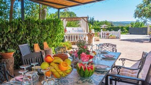 Un espace extérieur aménagé pour les repas, avec une table dressée pour deux personnes, entouré de verdure et un barbecue en arrière-plan.