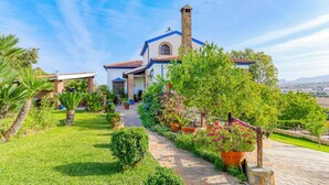 Une maison avec une cheminée en pierre, un toit en tuiles et un jardin où poussent différentes plantes, ainsi qu’un chemin menant à l’entrée.