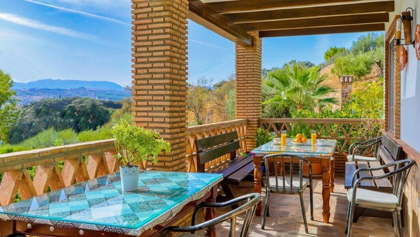 Une terrasse extérieure couverte, dotée d’une table et de chaises en verre, entourée de piliers en briques et d’une balustrade en bois.