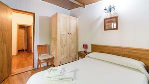 Una habitación con un armario de madera, una cama con ropa de cama blanca, una silla, una mesita de noche con una lámpara y un cuadro en la pared.