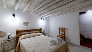 Quarto com cama de madeira, duas luminárias de cabeceira, uma cadeira e parede com textura rústica.