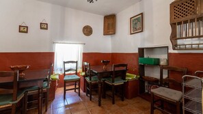 A dining area with wooden tables and chairs, a window with curtains, and a shelf with kitchenware. - Cortijo La Bodega - Conjunto Benaoján by Ruralidays (Málaga)
