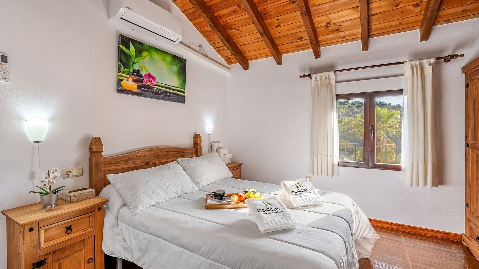 Une chambre avec un plafond en bois, un lit avec une literie blanche, une table de chevet avec une lampe, une fenêtre avec des rideaux et un tableau accroché au mur.