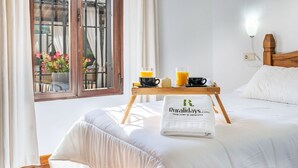 A neatly made bed with a white pillow and a folded towel, a wooden tray with breakfast items, and a window with curtains and flowers. - Iratxe Montefrío by Ruralidays (Granada)