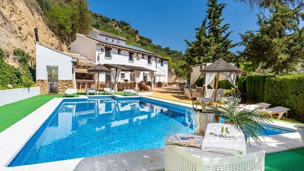 Une piscine avec une terrasse carrelée de carreaux blancs, un kiosque et un bâtiment à la façade en pierre.