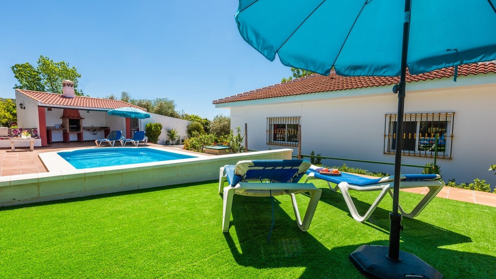 Une piscine avec une terrasse carrelée, entourée de gazon synthétique. On y trouve des chaises longues et un grand parasol qui offre de l’ombre.
