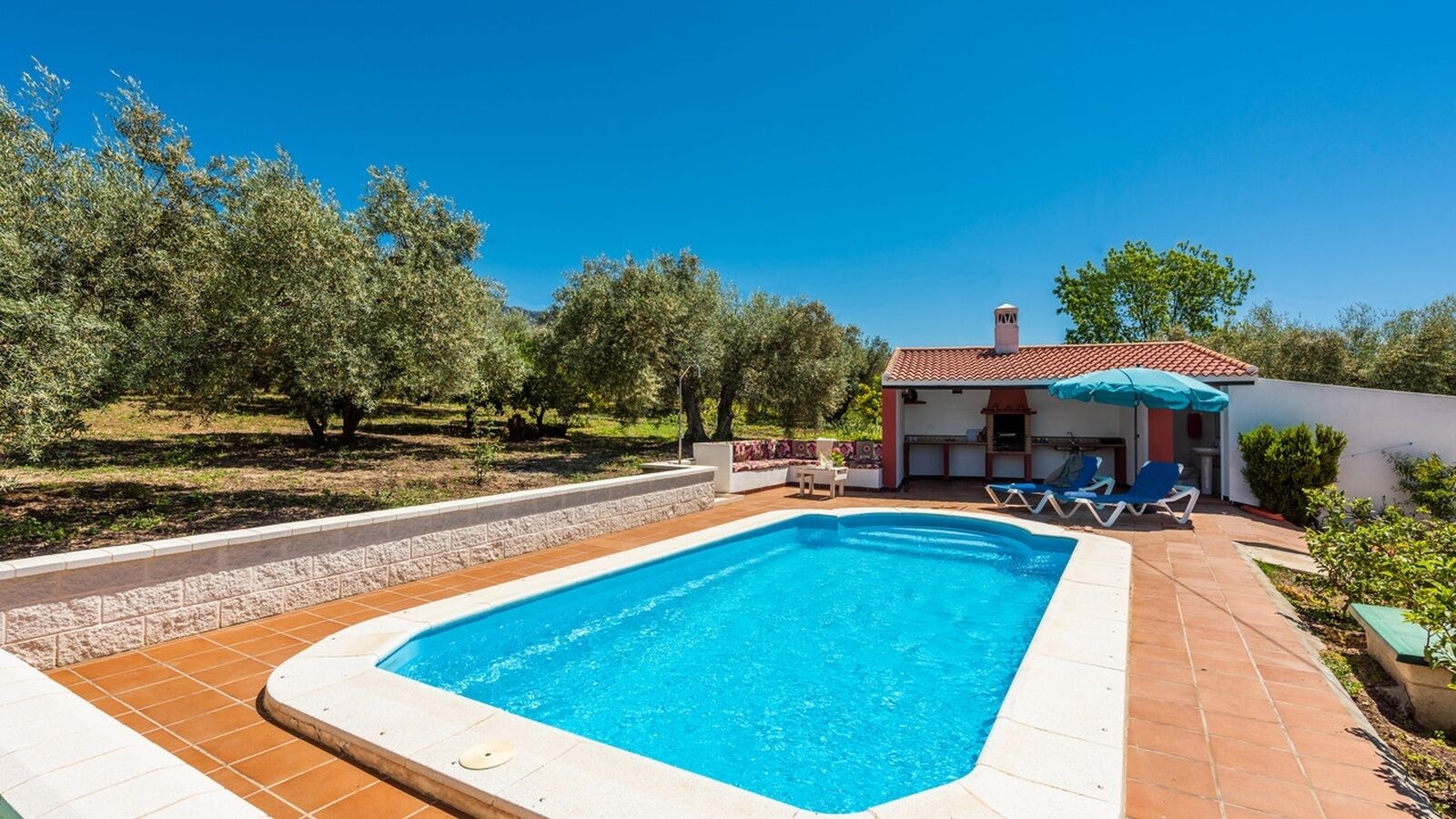A rectangular swimming pool with a tiled deck, surrounded by a low stone wall. There is a small building with a red-tiled roof and a blue umbrella in the background.