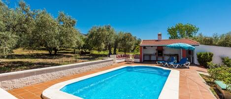 A rectangular swimming pool with a tiled deck, surrounded by a low stone wall. There is a small building with a red-tiled roof and a blue umbrella in the background.