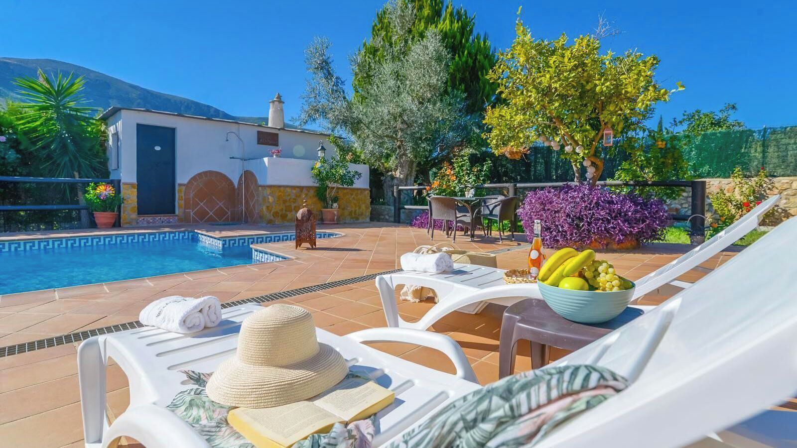 A poolside area with lounge chairs, a hat, a bowl of fruit, and a building with a tiled roof.