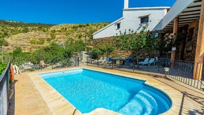 Uma piscina retangular com deck de pedra, cercada por uma grade de metal, e um edifício branco com detalhes em pedra ao fundo.