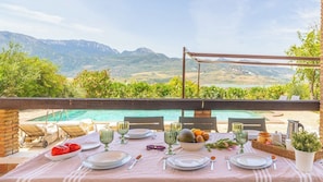 Mesa de jantar montada ao ar livre, vista para montanhas, piscina e cadeiras de descanso.