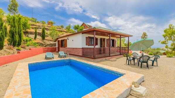 Un espace aménagé au bord de la piscine, avec une piscine rectangulaire, un patio couvert doté de mobilier d’extérieur et une colline en arrière-plan.