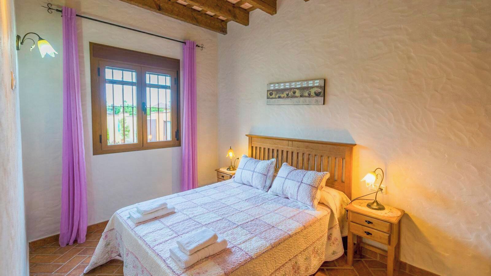 Un dormitorio con una cama, dos mesitas de noche, un ventanal con cortinas, una obra de arte en la pared y un techo de madera.