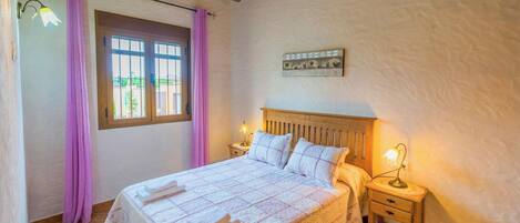 Ein Schlafzimmer mit einem Bett, zwei Nachttischen, einem Fenster mit Vorhängen, einem Wandbild und einer Holzbalkendecke.