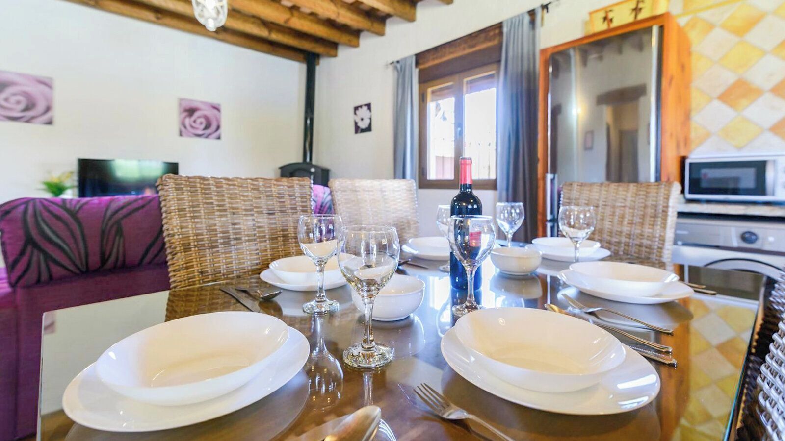 Una mesa de comedor dispuesta con platos, vasos y cubiertos, rodeada de sillas de mimbre.
