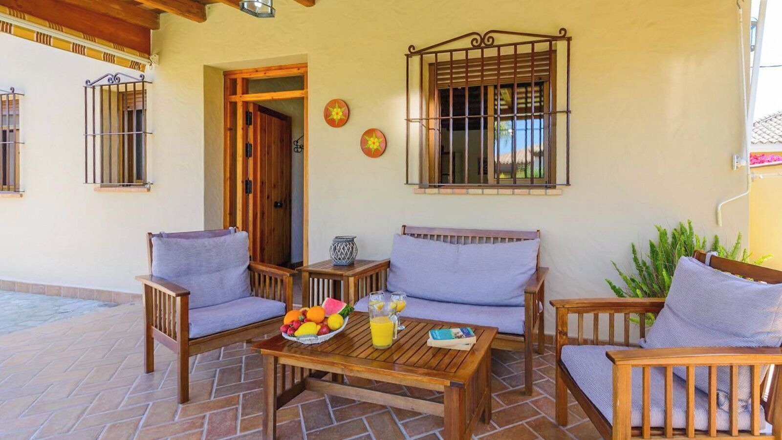 A patio with wooden furniture, a table with fruit and drinks, and a window with bars.