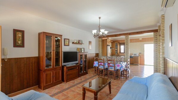 Ein geräumiges Wohnzimmer mit Fernseher, Holzelementen, einem Essbereich mit Tisch und Stühlen sowie einer Küche im Hintergrund.