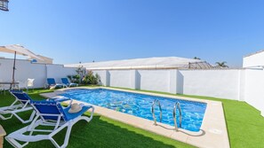 Ein rechteckiger Swimmingpool mit klarem, blauem Wasser, umgeben von künstlichem grünen Rasen und weißen Wänden. Blaue Liegestühle und ein weißer Sonnenschirm sind vorhanden.