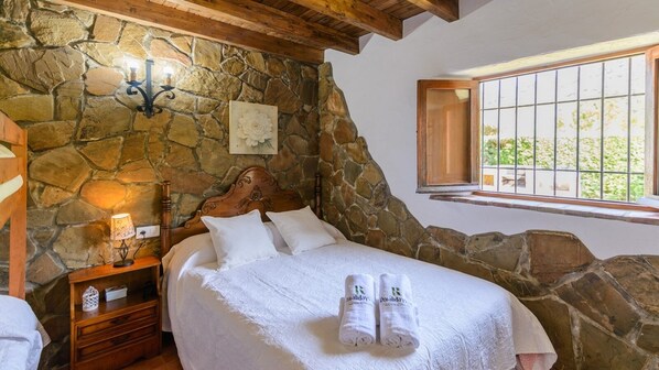 A bedroom with a stone wall, a wooden headboard, a nightstand with a lamp, and a window with bars. - Los Cascajales Benaoján by Ruralidays (Málaga)