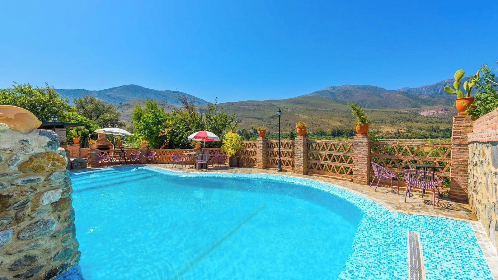 A swimming pool with a stone wall and chairs, surrounded by a fence and potted plants, with a mountainous landscape in the background.
