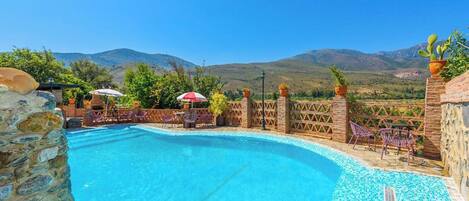 A swimming pool with a stone wall and chairs, surrounded by a fence and potted plants, with a mountainous landscape in the background.