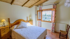 A bedroom with a wooden bed, a nightstand with a lamp, a chair, a window with curtains, and a ceiling fan. - Cortijo San Jose Iznájar by Ruralidays (Córdoba)