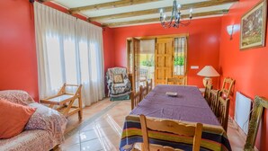 A room with a dining table, chairs, a sofa, a chair with a cushion, a chandelier, and a door leading outside. - Cortijo San Jose Iznájar by Ruralidays (Córdoba)