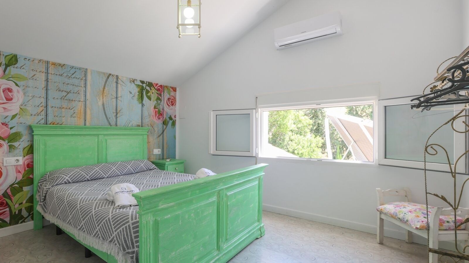 Un dormitorio con una cama verde, una mesita de noche, una silla, un ventanal y una pared con papel tapiz floral.