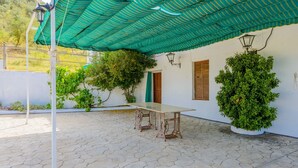 Un patio al aire libre techado, con una mesa y sillas, rodeado de follaje y un edificio blanco.