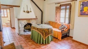 Una habitación acogedora con chimenea de piedra, una mesa redonda con mantel verde, un sofá a rayas y una ventana con cortinas.