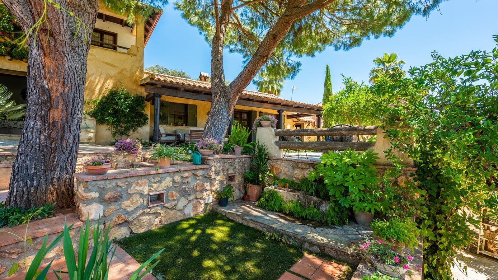 A Mediterranean-style villa with stone steps leading to a patio, surrounded by lush greenery and large trees.
