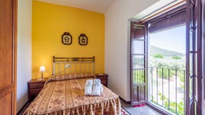 A bedroom with a bed, two wall-mounted clocks, a bedside lamp, and a balcony with a view of greenery.