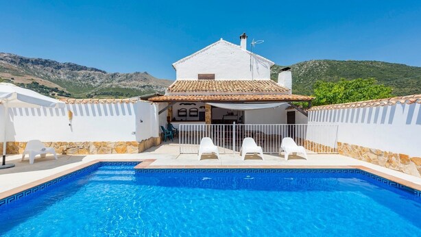 A swimming pool with a white house in the background, surrounded by stone walls and a mountainous landscape.