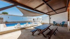 A covered outdoor area with a pool, white lounge chairs, and a wooden table.