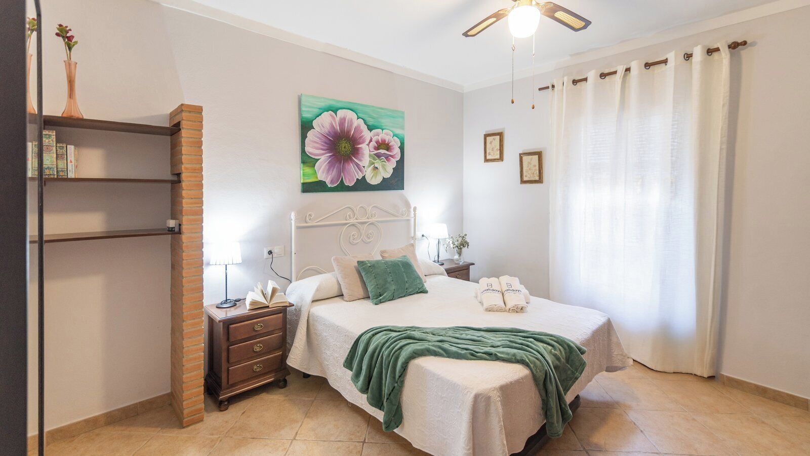 A bedroom with a bed, bedside tables, a nightstand, a bookshelf, a painting, and a window with curtains.