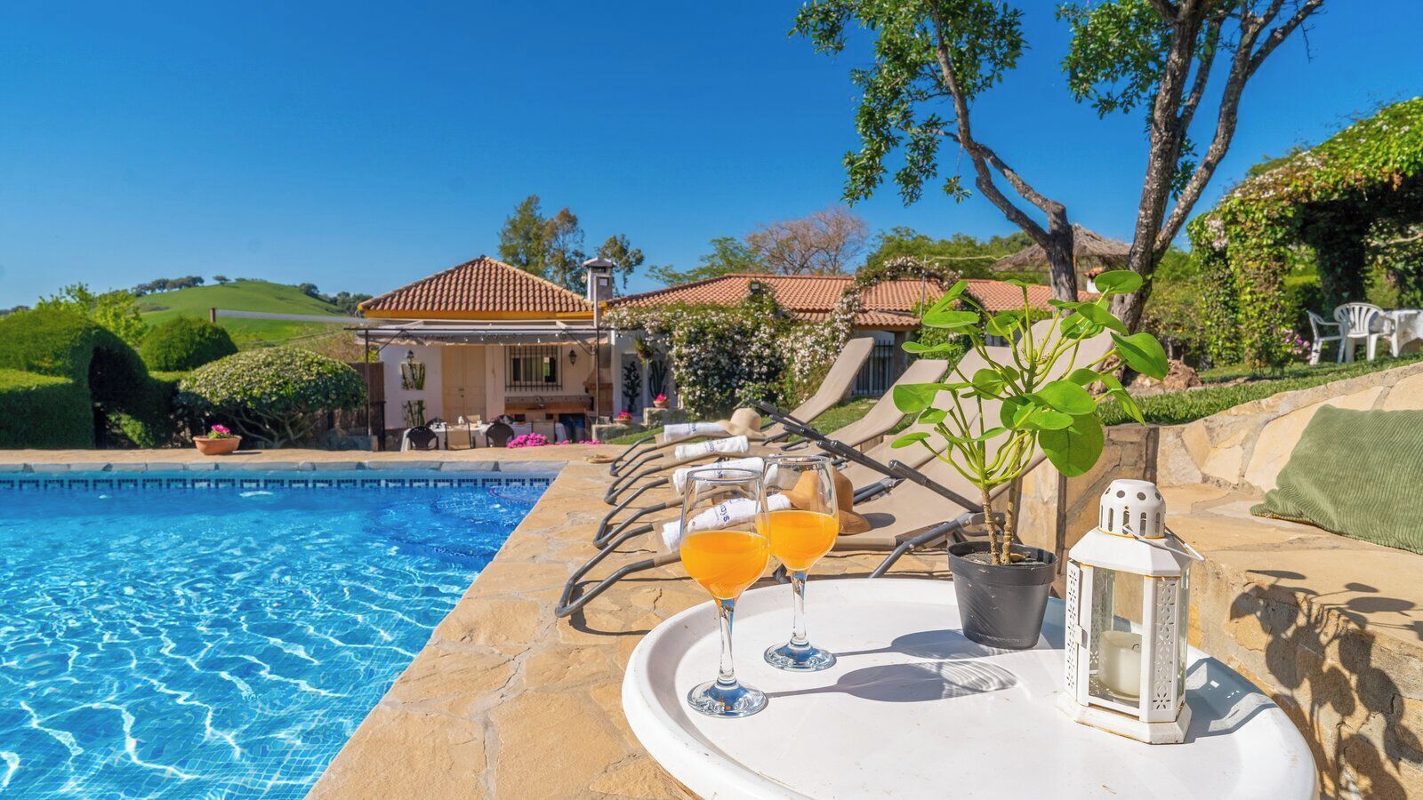 A poolside area with a clear blue pool, lounge chairs, and a table with two glasses of orange juice.