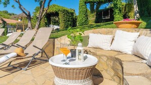 Un patio de piedra con sillones de descanso, una mesa de mimbre con bebidas y una zona de estar acolchada.