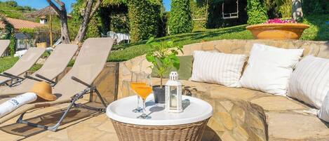 Un patio de piedra con sillones de descanso, una mesa de mimbre con bebidas y una zona de estar acolchada.