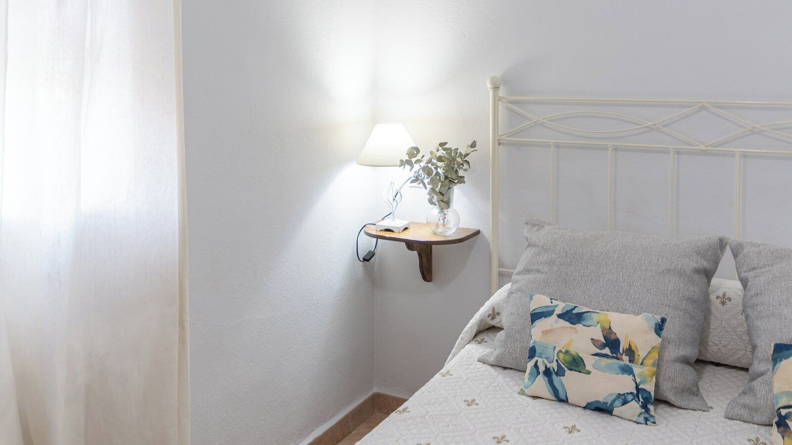 Un dormitorio con cama, una mesita de noche con lámpara y un florero, y cabecera blanca.